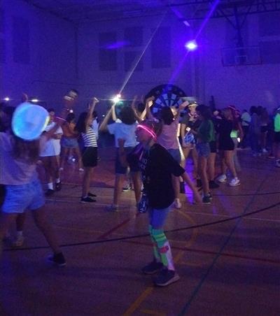 A crowd of middle school aged kids dancing in a basketball gym, waving phone flashlights with the overhead lights off and blacklights on while dancing.