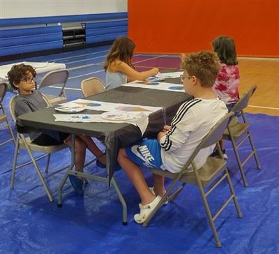 4 children sit around a table, there are papers with painted ice cream cone pictures on them and one child is actively painting.