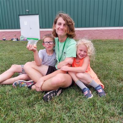 A camp counselor sits in the grass with a child on each side clinging to his arms, all smiling.