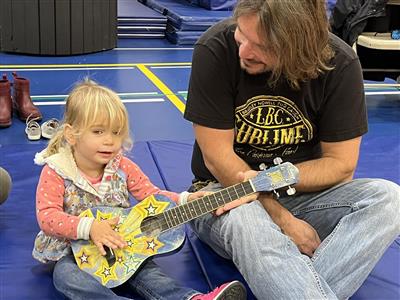 An adult sits on a gymnastics mat on the floor beside a toddler holding a ukulele.