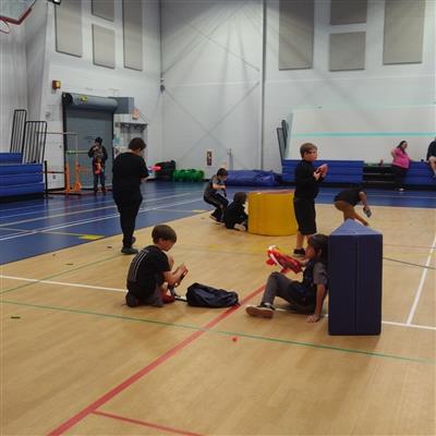 Children crouched behind gymnastics mat obstacles in a basketball gym holding nerf blasters with nerf darts scattered across the floor.